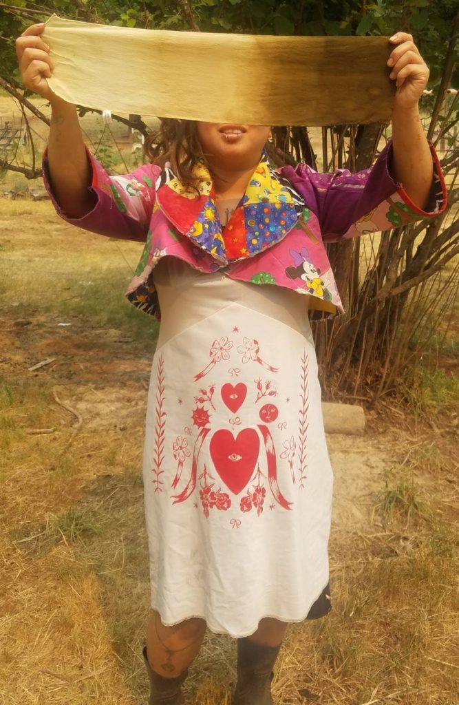 R. Silver stands outside, holding a naturally dyed piece of silk in front of her eyes. She is wearing a quilted bolero and white slip dress with a screen print by Serpent and Bow