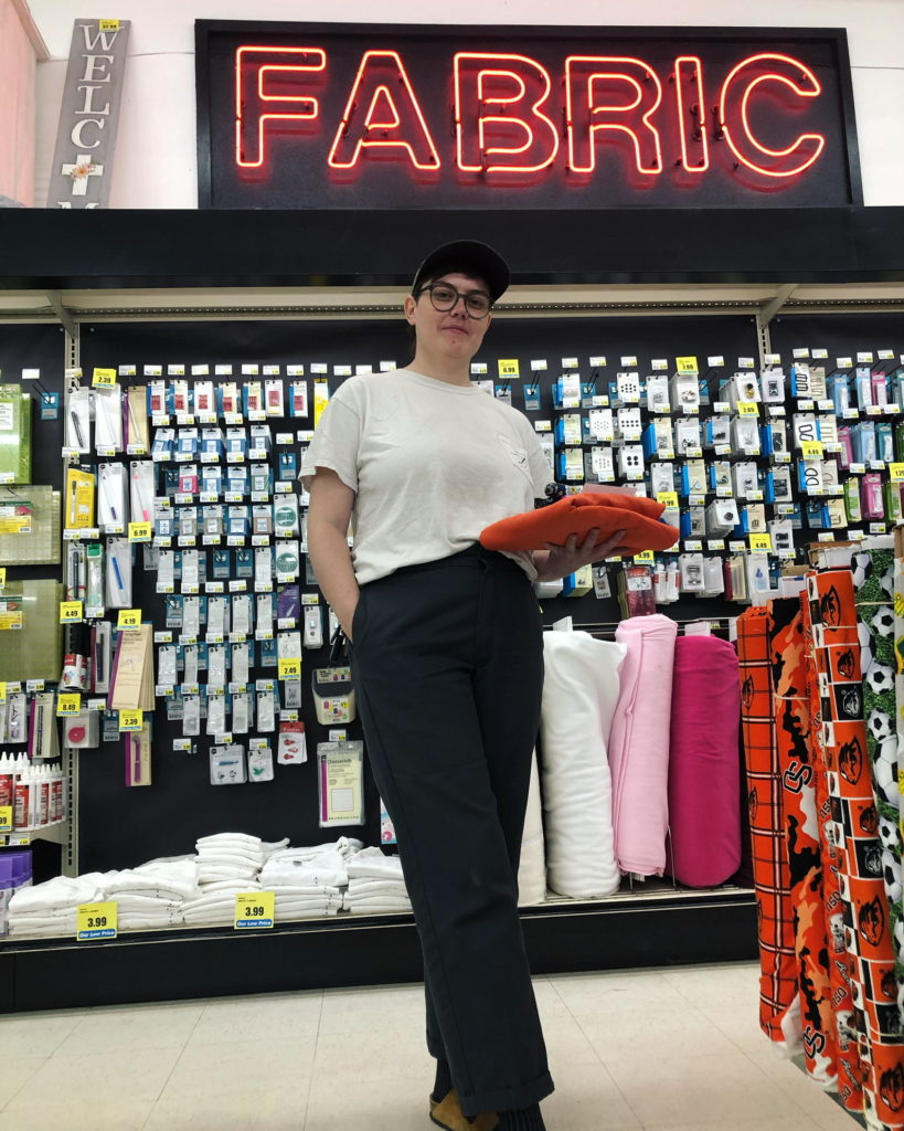 GK Bigler stands in a fabric store. wearing a white tshirt and black plants in front of a notions wall.