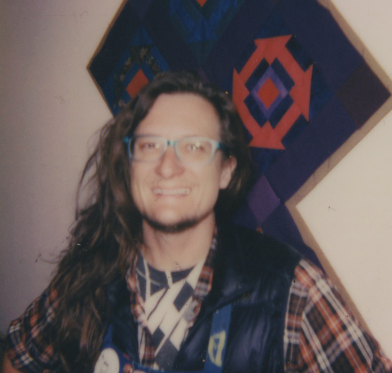 Headshot of Ian. He is wearing a blue vest over a plaid shirt. There is a 4-square blue quilt wall hanging he behind him.