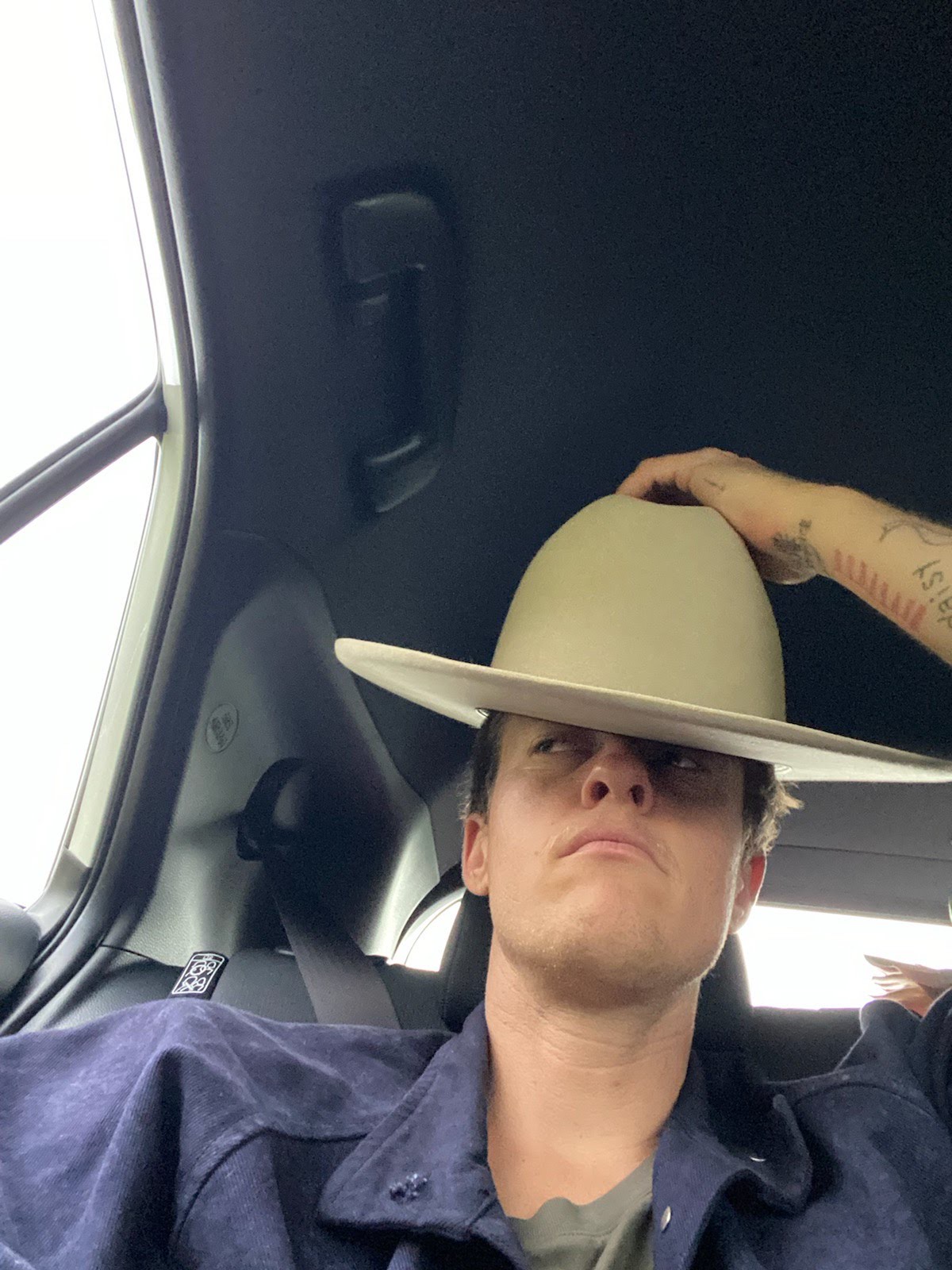 Laub sits in a car with a cowboy hat draped tilted to the right, almost covering one eye.  He glares to the left. His left hand rests on his hat.