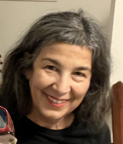 George smiles at the camera in front of a white wall. She is wearing a black shirt. She has shoulder length salt and pepper hair with baby bangs.